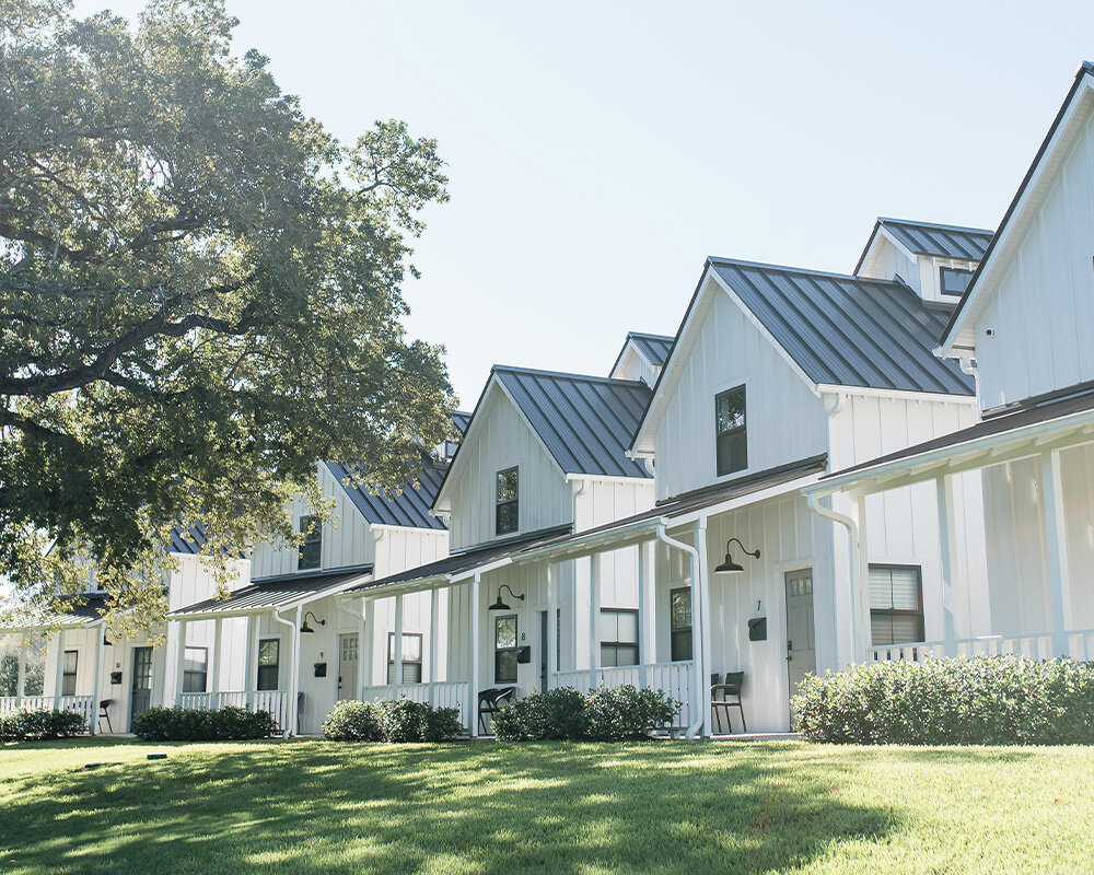 A row of modern white farmhouse-style cabins with metal roofs and covered front porches in a sunny, grassy field.