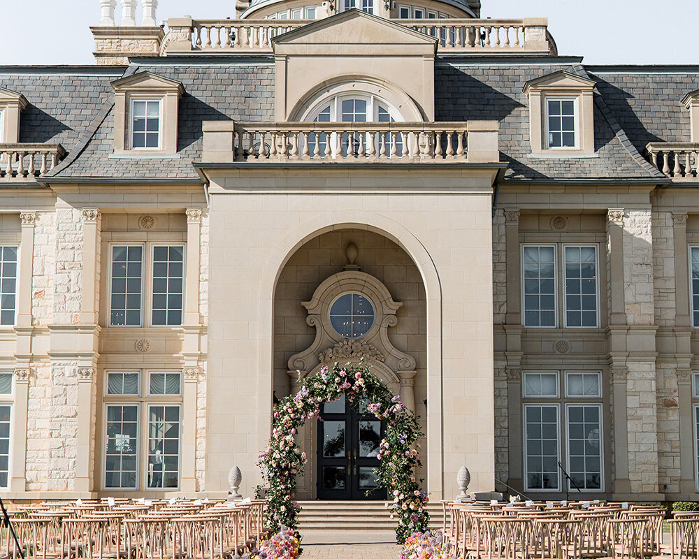 The exterior of a luxury stone estate wedding venue featuring a grand entrance arch decorated with a floral archway and an outdoor ceremony setup with rows of wooden chairs.