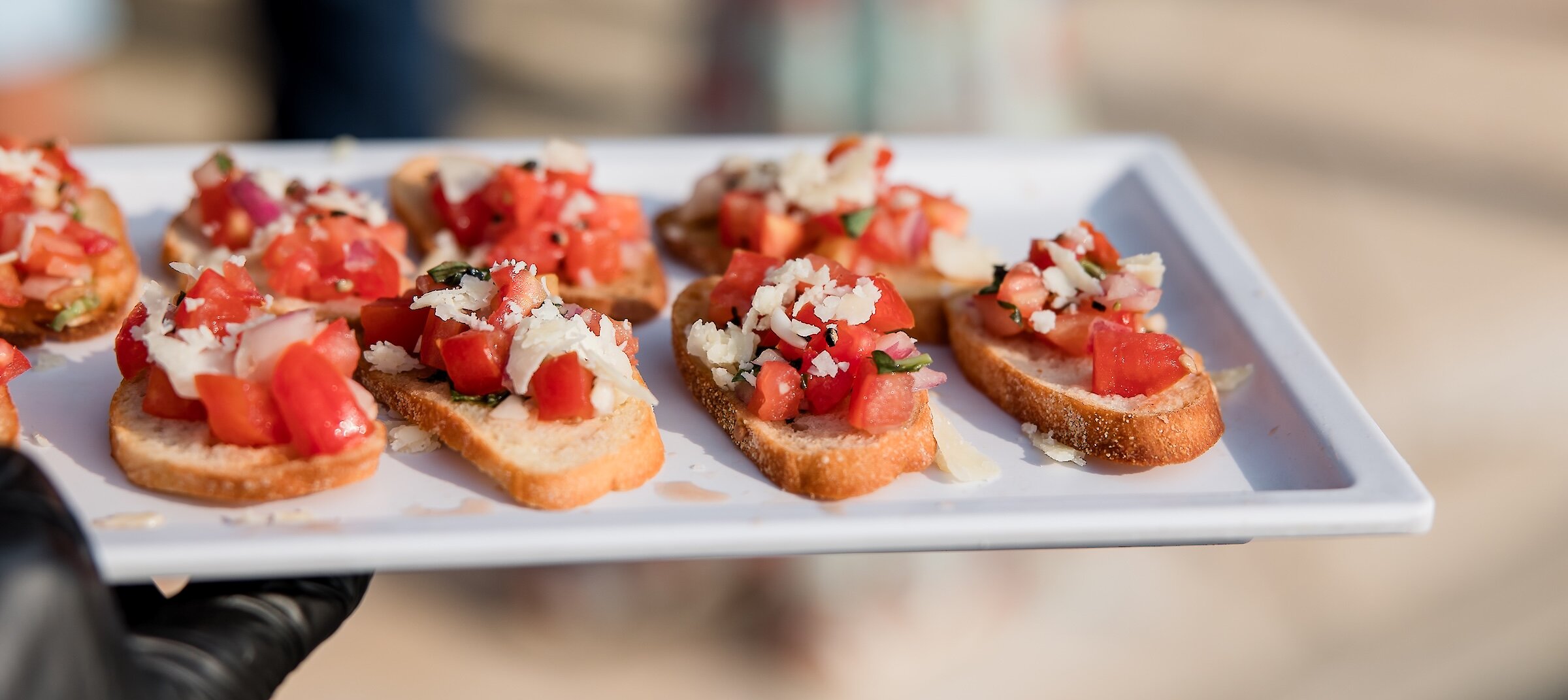 A server wearing a black glove holds a white tray of fresh bruschetta appetizers topped with diced tomatoes and grated cheese.