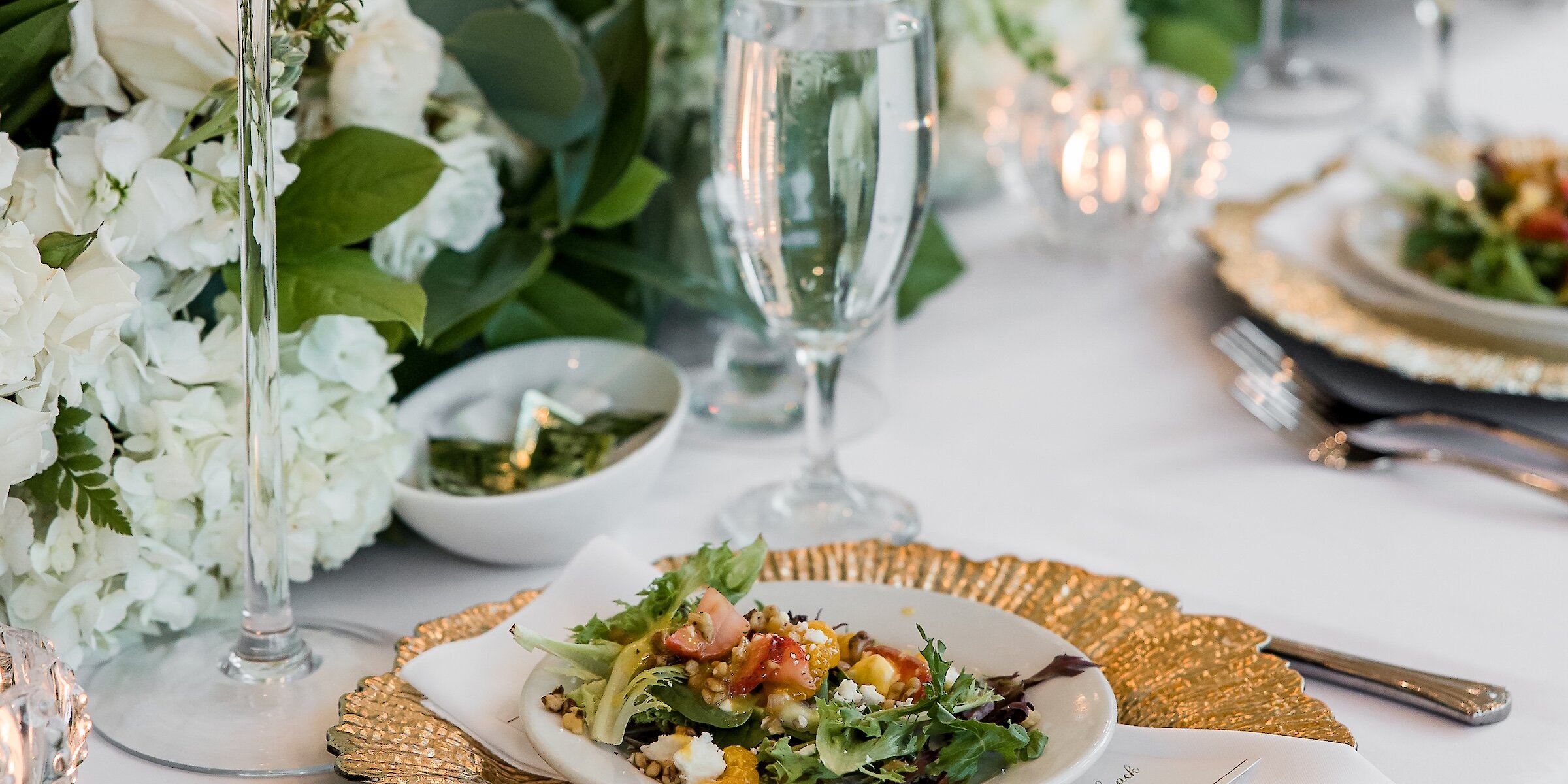 A wedding table detail featuring a plated salad on a gold ruffled charger plate, surrounded by white floral centerpieces and candles.