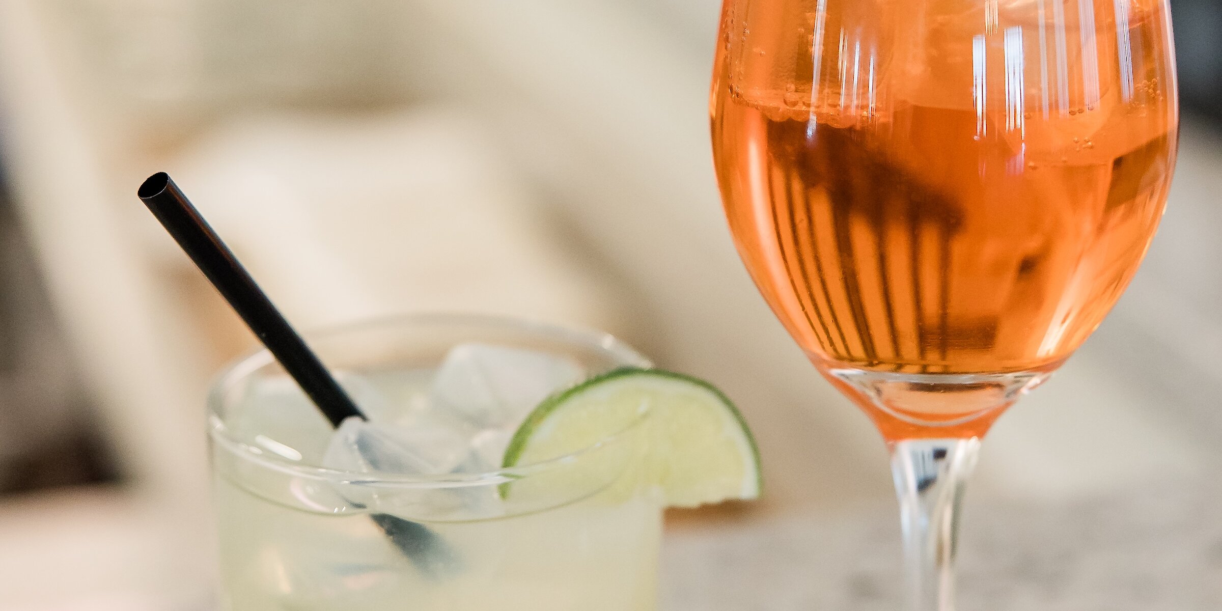 A close-up of two wedding cocktails: a light yellow drink with a lime wedge and a bright orange spritz in a wine glass.
