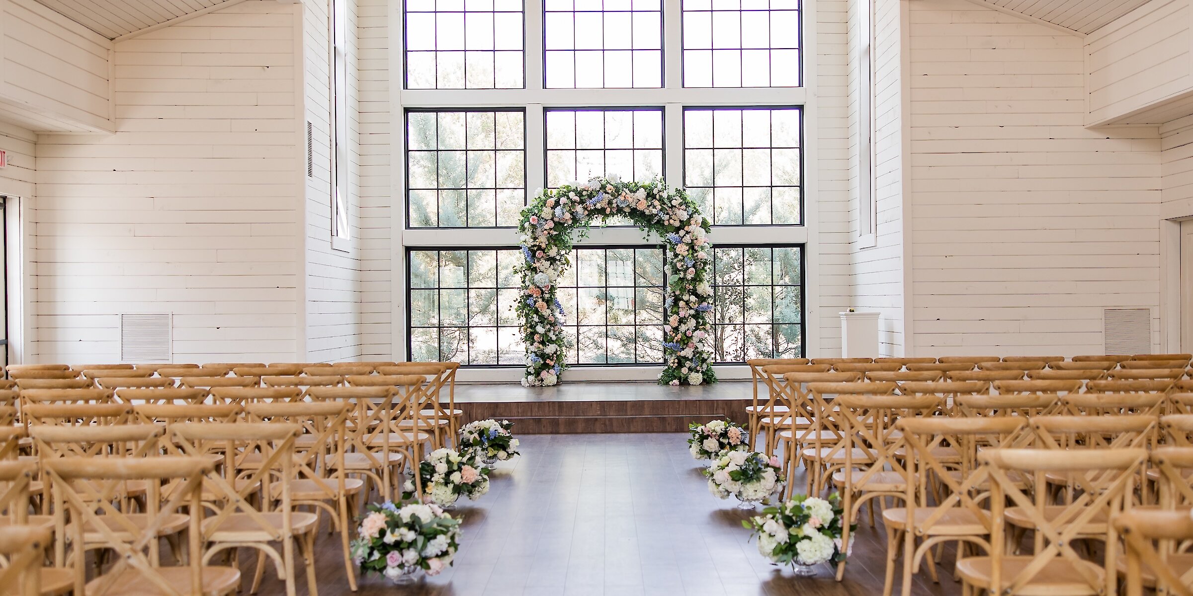 The bright interior of a farmhouse wedding chapel with white shiplap walls, wood floors, rows of cross-back chairs, and a floral arch.
