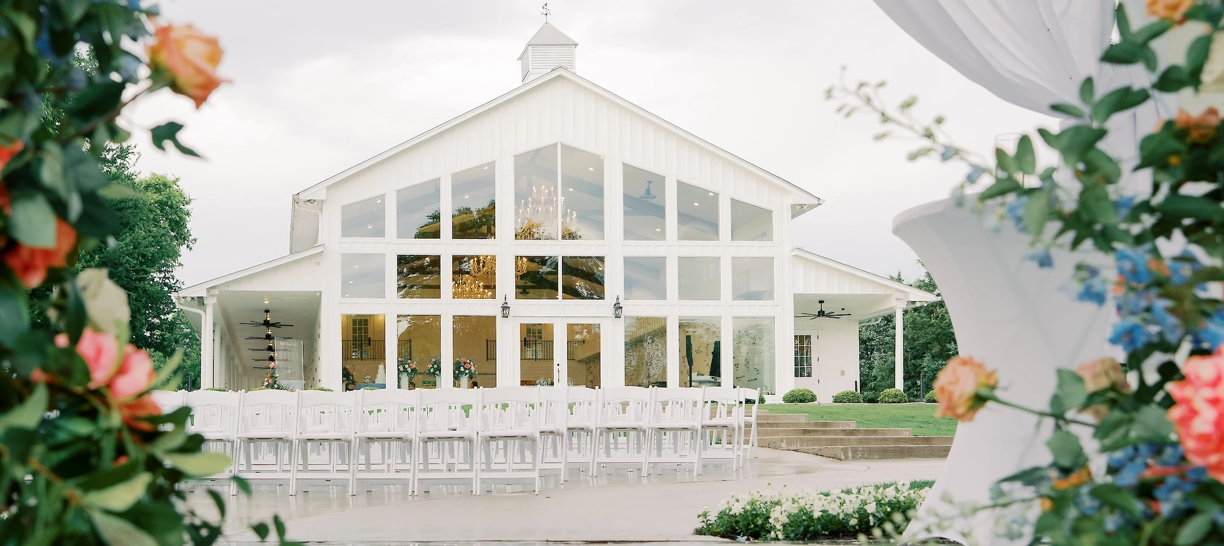 An outdoor wedding ceremony site with white chairs facing a modern white barn venue with large glass windows, framed by sheer fabric and flowers.