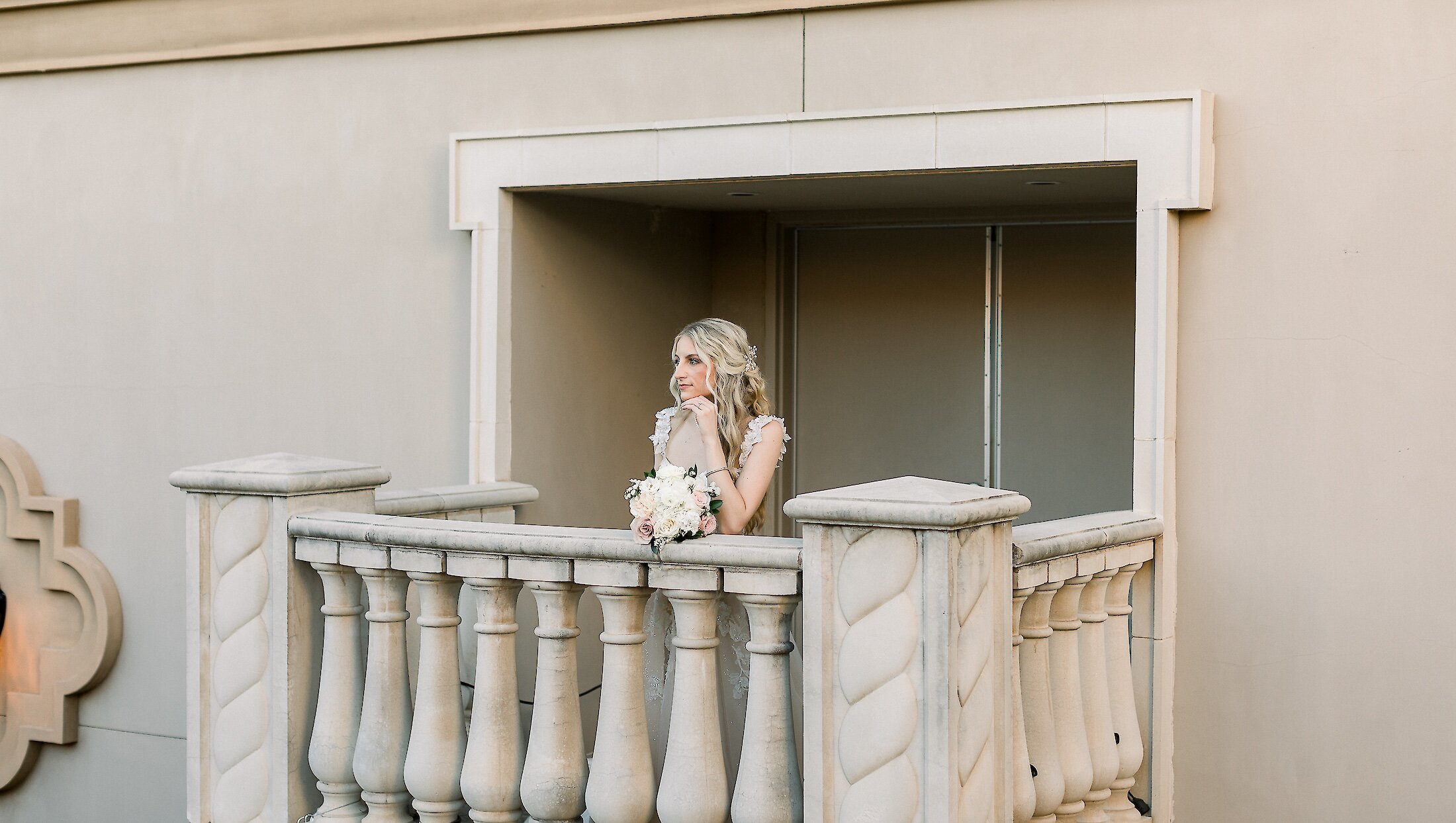 A beautiful blonde bride in her gown is standing on a balcony with a stone balustrade, holding a white and pink bouquet.