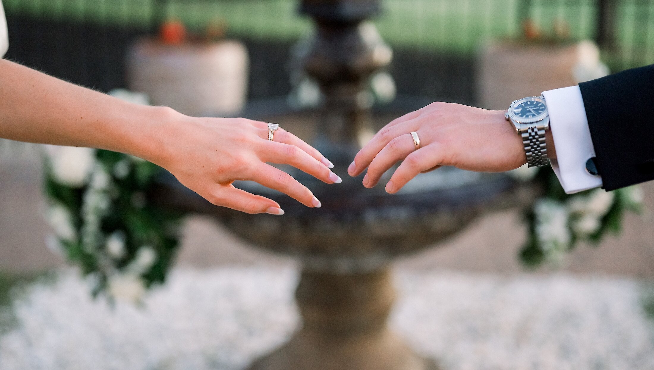 Close-up photo of the bride and groom's hands reaching toward each other, displaying the engagement ring and wedding band over a blurry fountain backdrop.