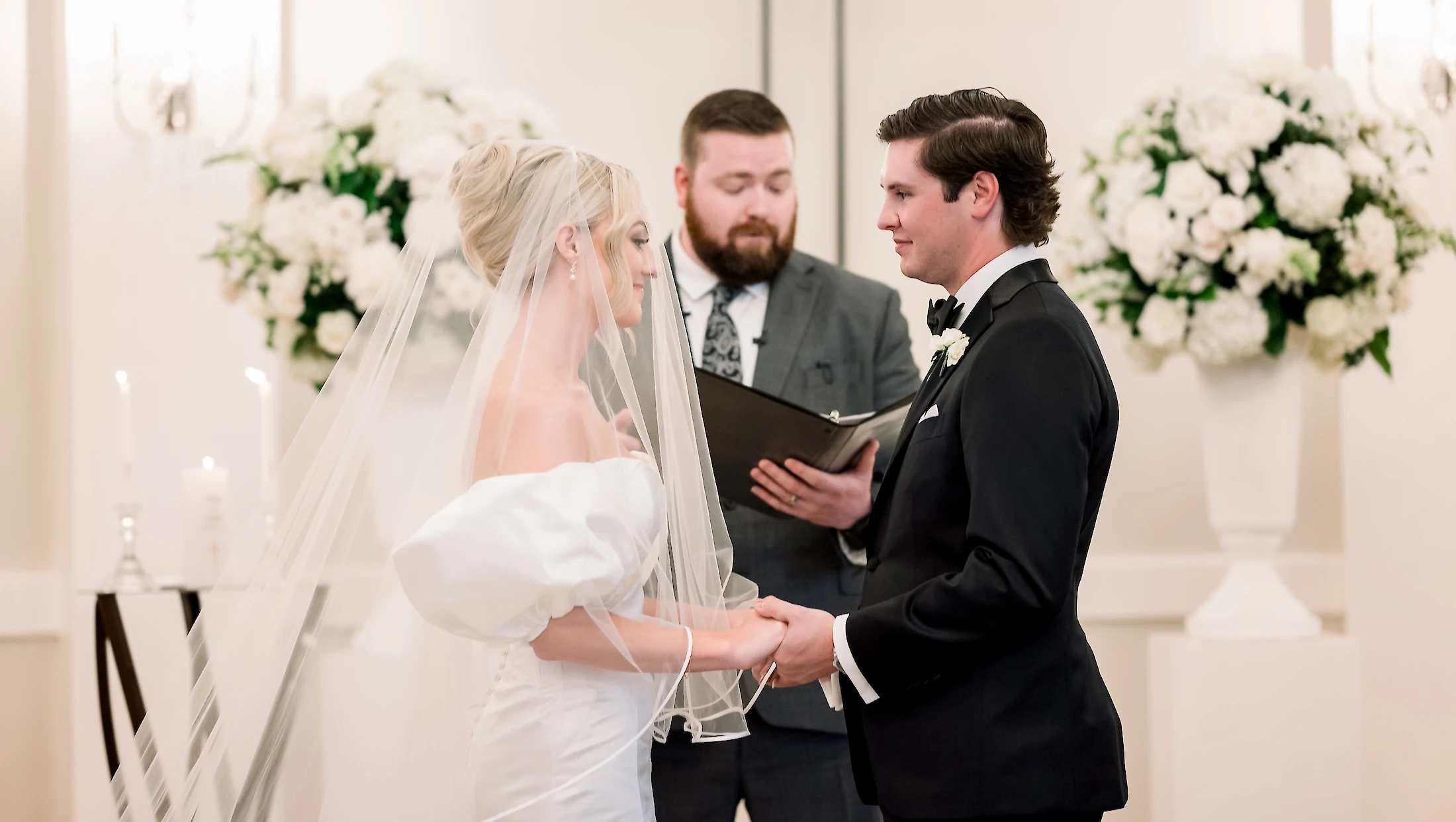 The bride and groom hold hands and face each other during their indoor wedding ceremony, with the officiant reading from a book between two large white floral arrangements.