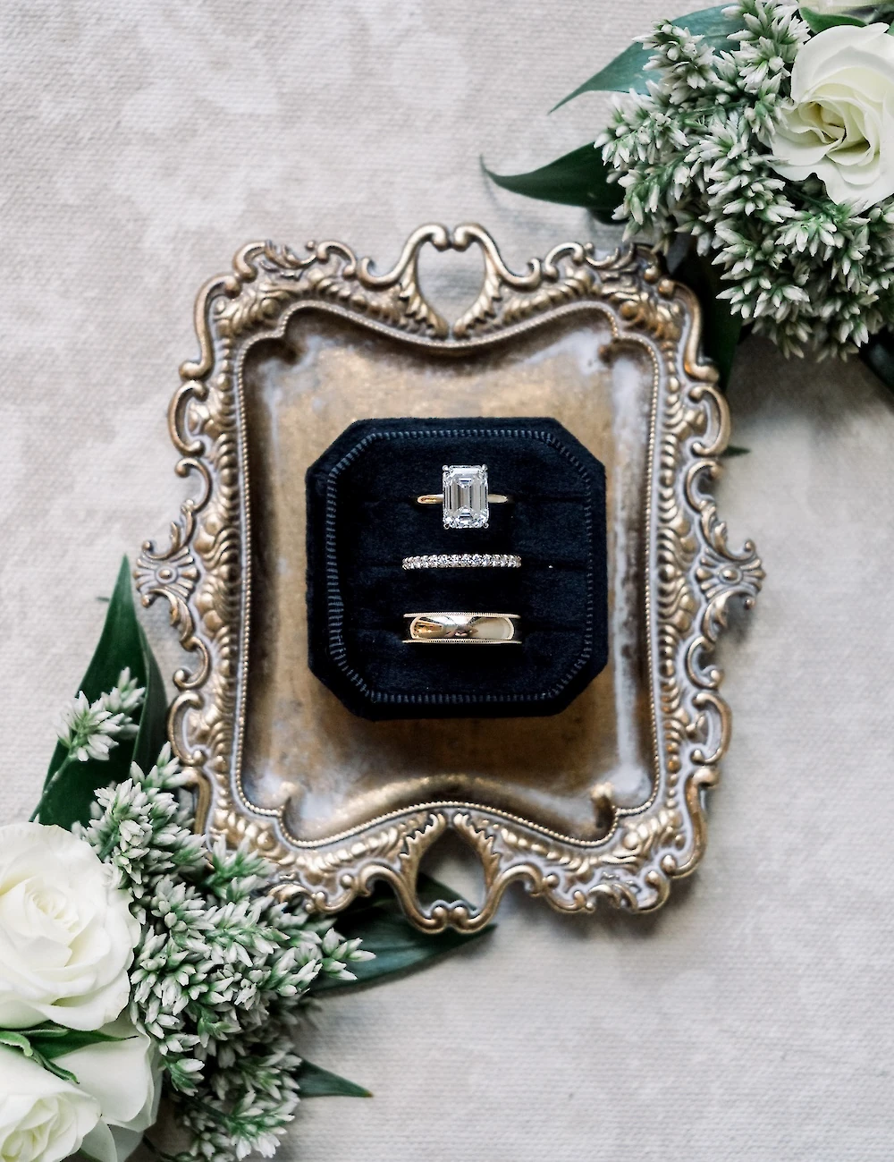 Close-up of three wedding rings, including a solitaire emerald-cut diamond ring, displayed in a black velvet box on a decorative gold antique tray with white roses.