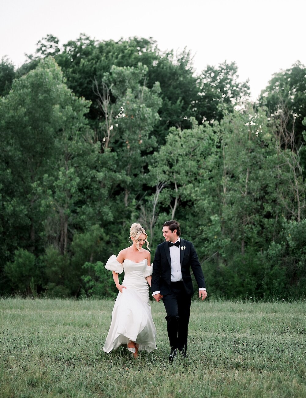 The bride and groom smile at each other while walking hand-in-hand through a grassy field with a lush backdrop of dark green trees.