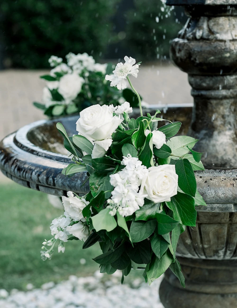 A wedding floral arrangement featuring white roses and greenery is draped over the edge of a tiered stone fountain outdoors.