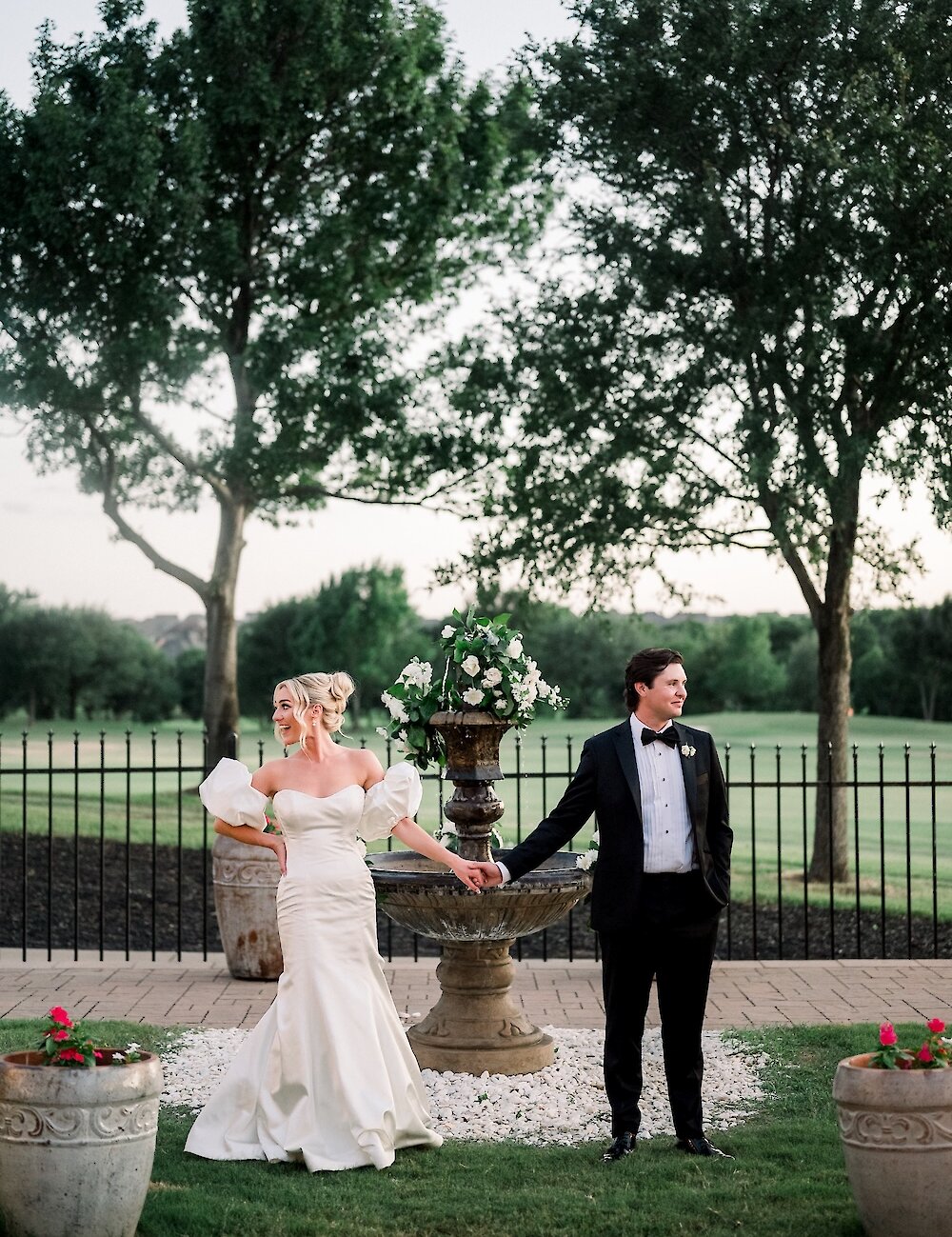 The bride and groom hold hands on opposite sides of a tiered floral fountain for an outdoor portrait against a backdrop of trees and a black fence.