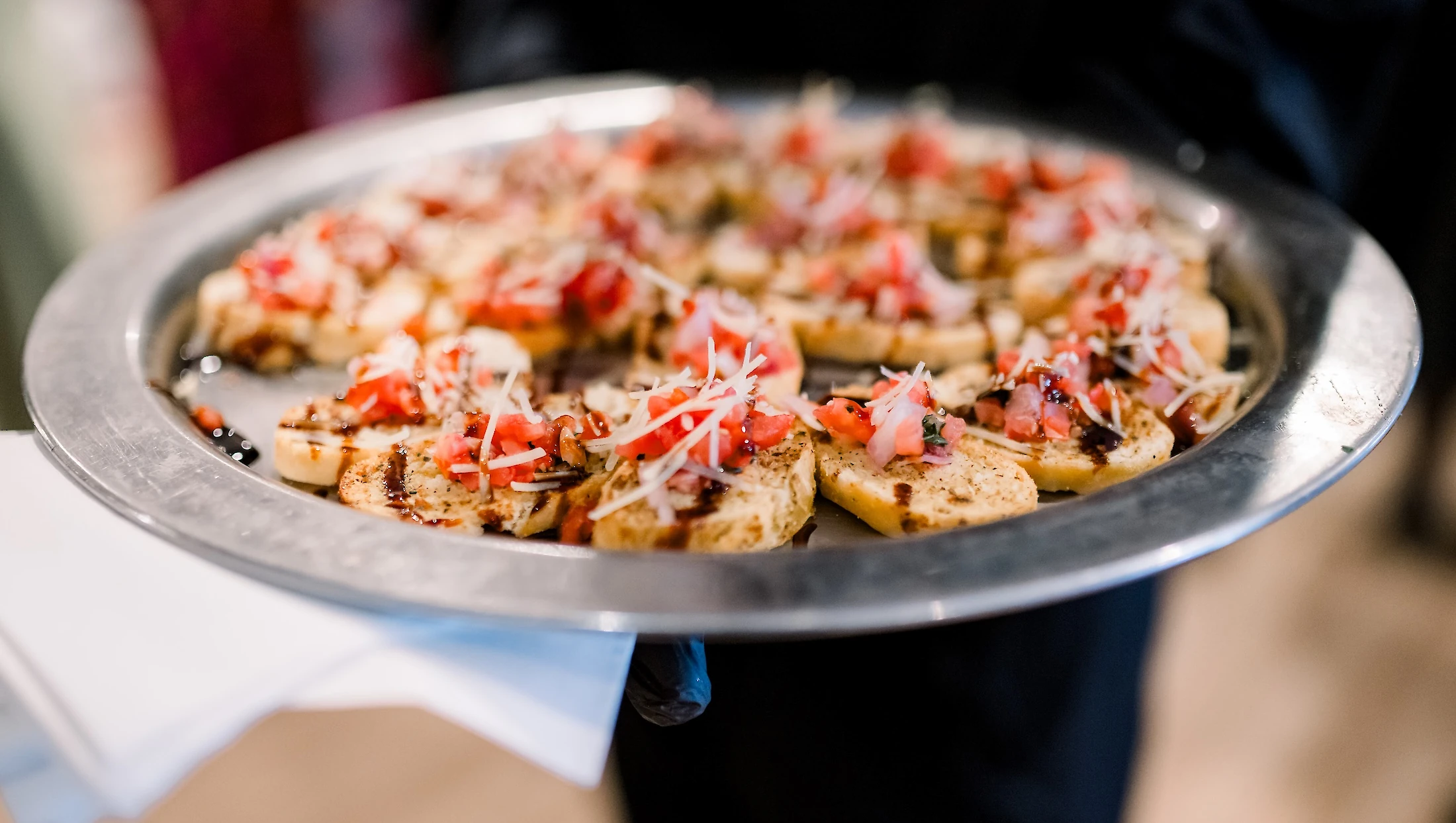 A caterer holds a silver platter of toasted bread appetizers topped with diced tomatoes, cheese, and a balsamic glaze.