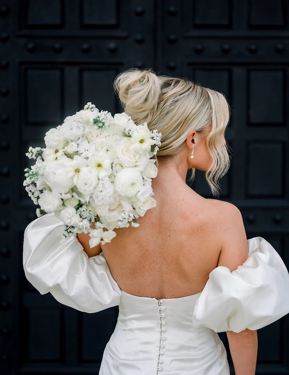 Rear view of the bride holding a large white bouquet, showcasing her off-the-shoulder dress with a row of buttons and her blonde hair styled in an updo.