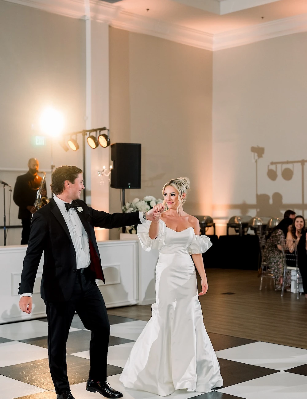 The bride and groom smile while dancing their first dance on a black and white checkered floor in a brightly lit reception ballroom.