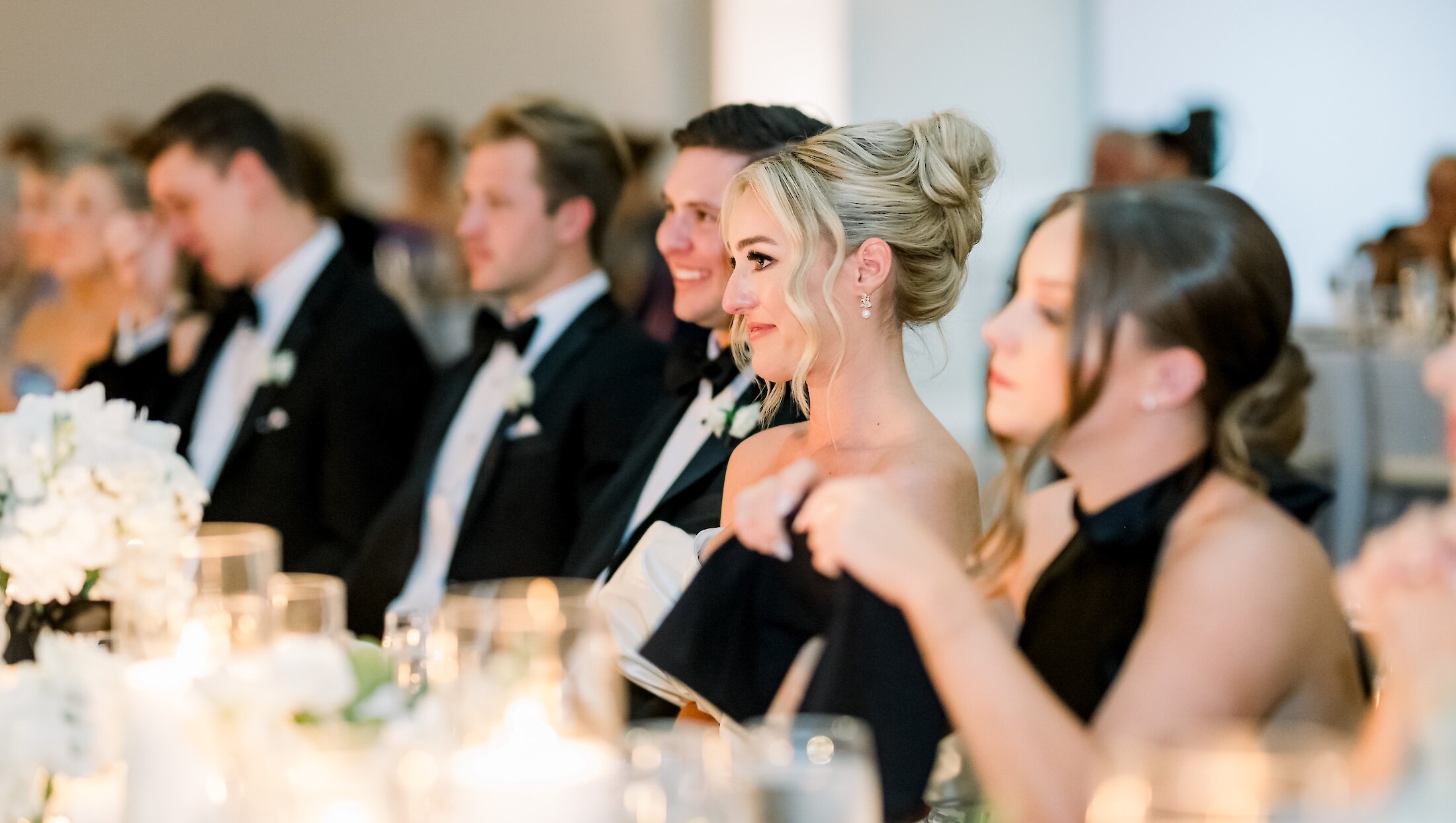 The smiling bride and the wedding party sit at the head table, with the table covered in white florals and glowing candlelight during the reception.