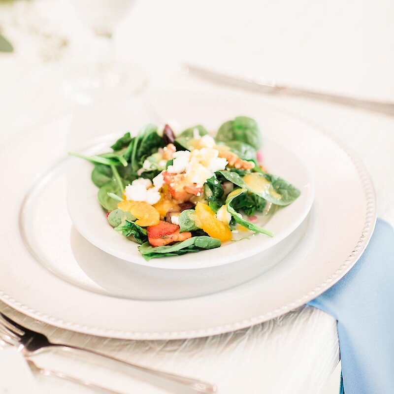 A small, elegant plated salad featuring spinach, orange segments, and feta cheese on a white charger plate at a wedding reception.