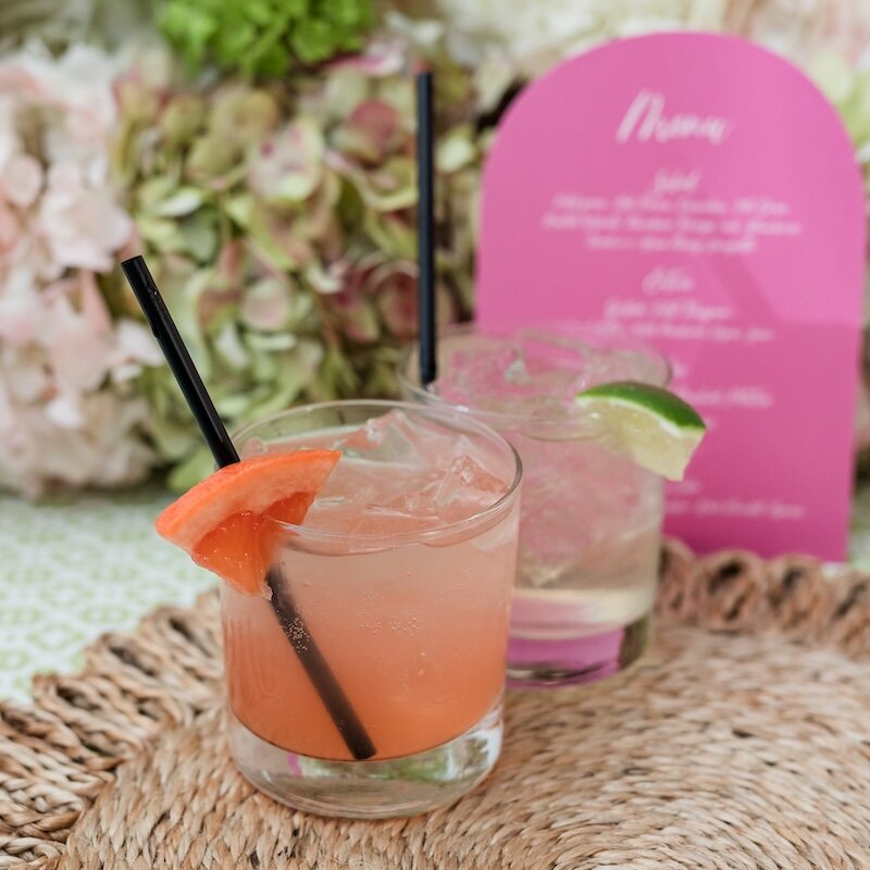 Two colorful cocktails served on a woven placemat, with a pink arched menu and a blurred floral background.