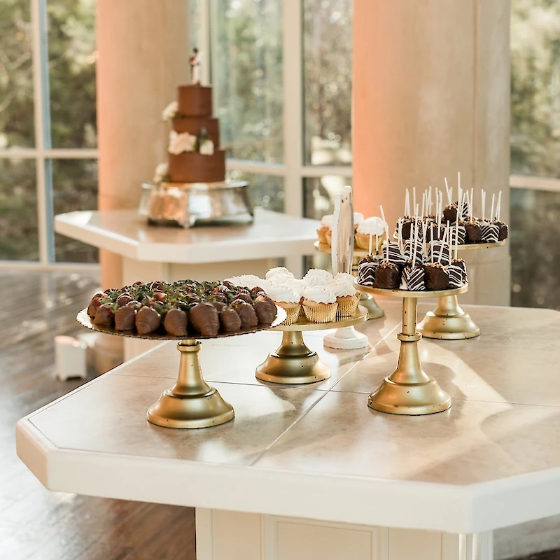 A wedding dessert table featuring chocolate-covered strawberries, cupcakes, and cake pops on gold pedestal stands with a tiered chocolate cake in the background.