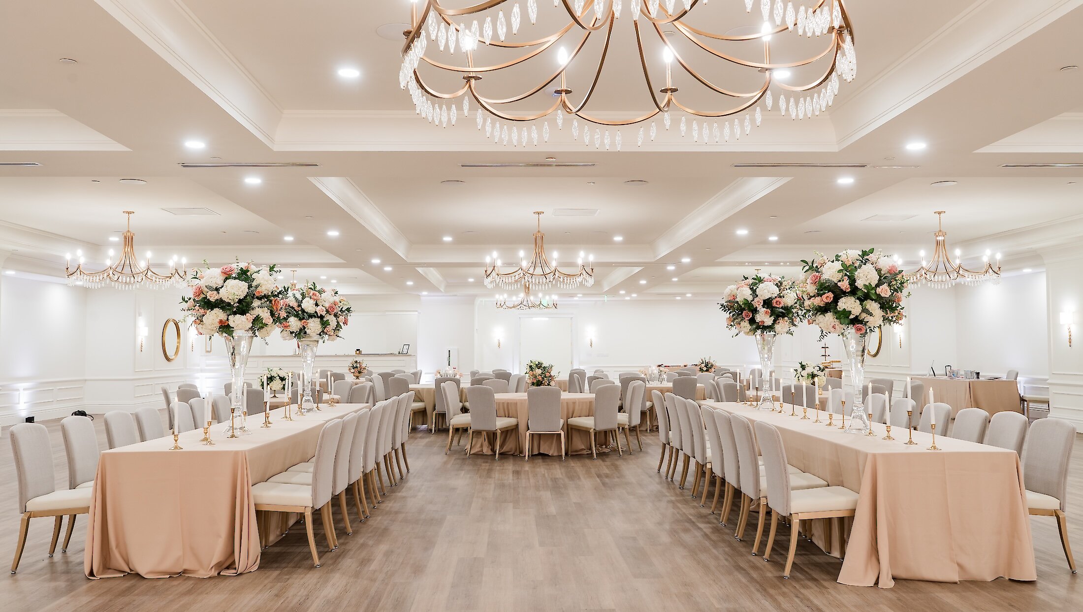 An elegant ballroom reception setup featuring long rectangular tables and round tables draped in blush pink linens, illuminated by multiple gold chandeliers and two grand floral centerpieces.