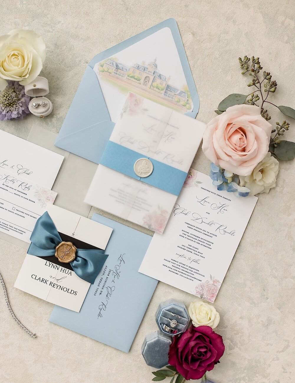 A formal wedding invitation flat lay featuring light blue envelopes, a white vellum wrap secured with a wax seal, and accents of pink and burgundy roses.