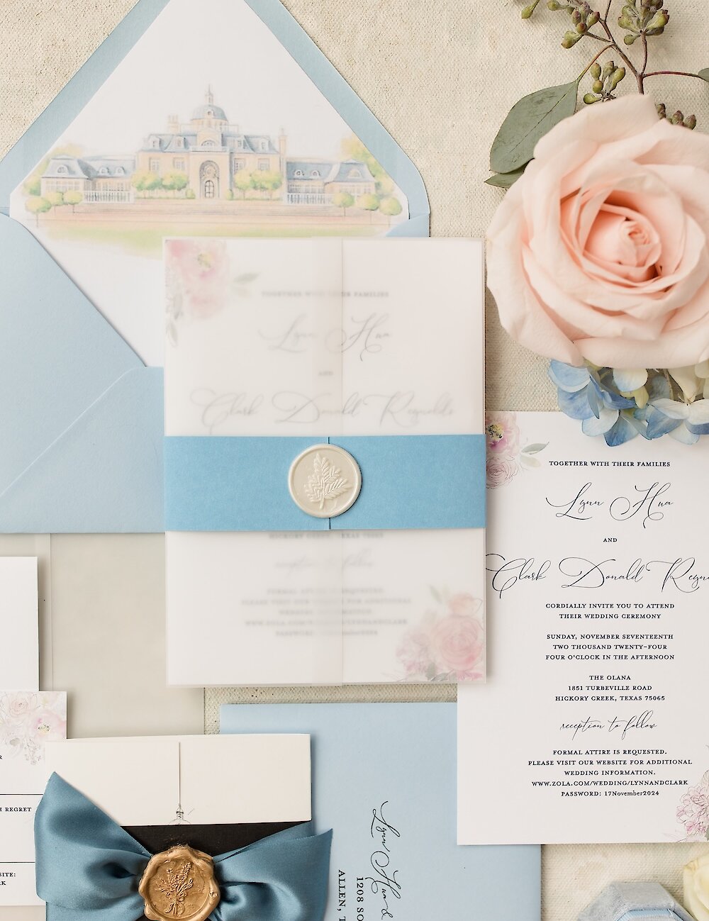 Close-up of a wedding invitation set with a translucent vellum wrap, a light blue belly band sealed with a white wax seal, and a blush pink rose detail.
