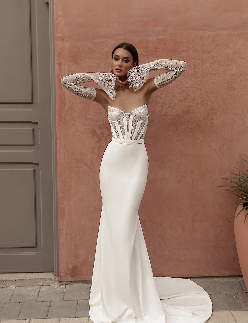 A model wearing a white fitted wedding gown featuring a sheer corset bodice and separate long lace sleeves with flared cuffs, posing against a terracotta wall.