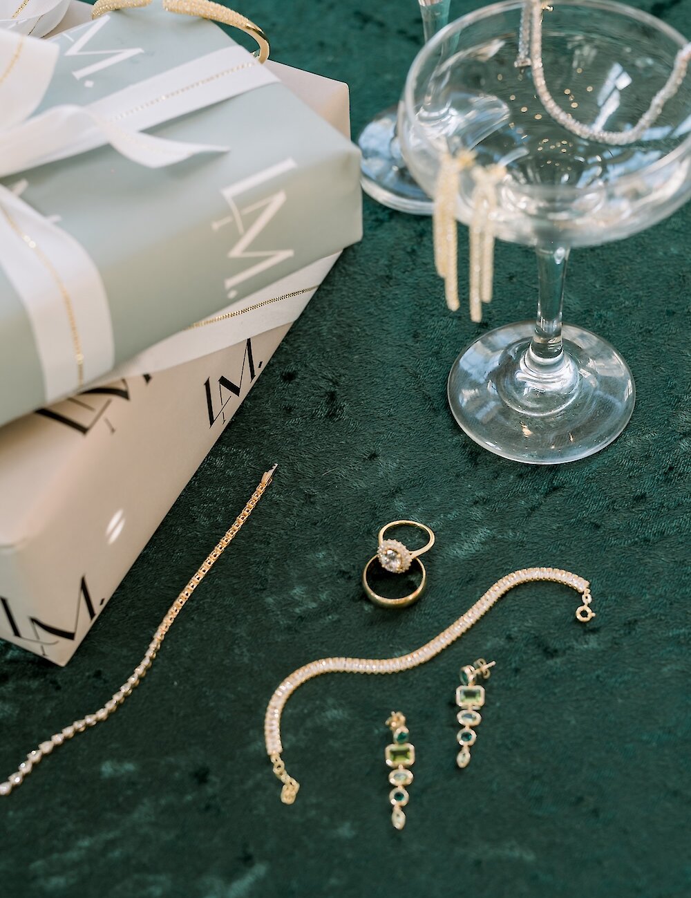 A flat lay of luxury bridal jewelry including diamond rings, gold tennis bracelets, and green emerald earrings, displayed on a dark green surface next to wrapped gifts.
