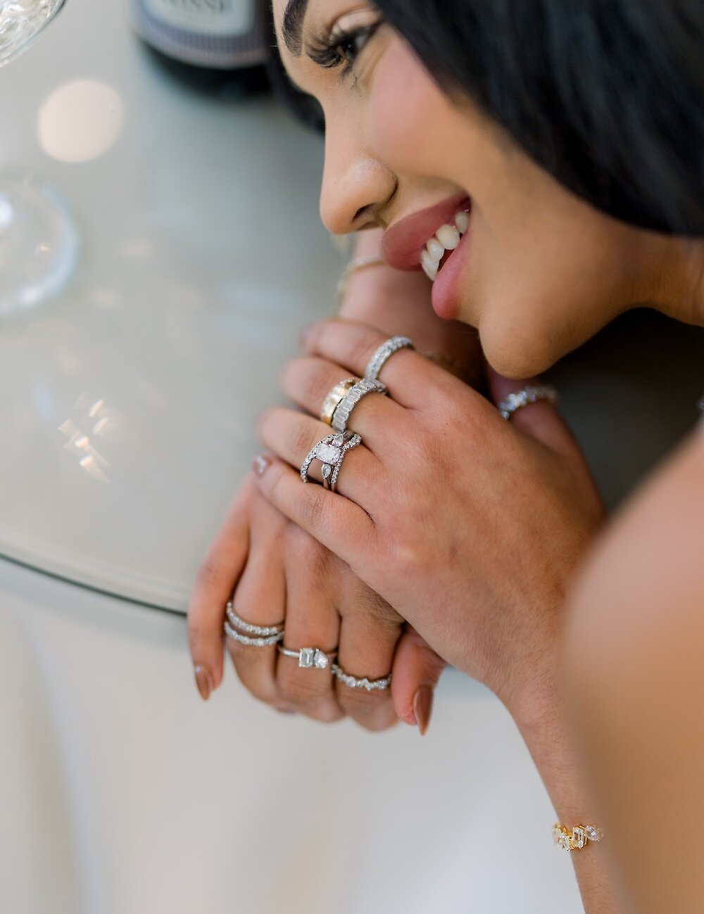 A close-up of a smiling bride model showing off multiple diamond rings, including a large engagement ring, on both hands and a diamond bracelet.