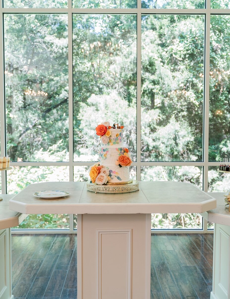 A tiered wedding cake decorated with orange and pink flowers is displayed on a dessert table with cookies and treats in a sunlit room with floor-to-ceiling windows.
