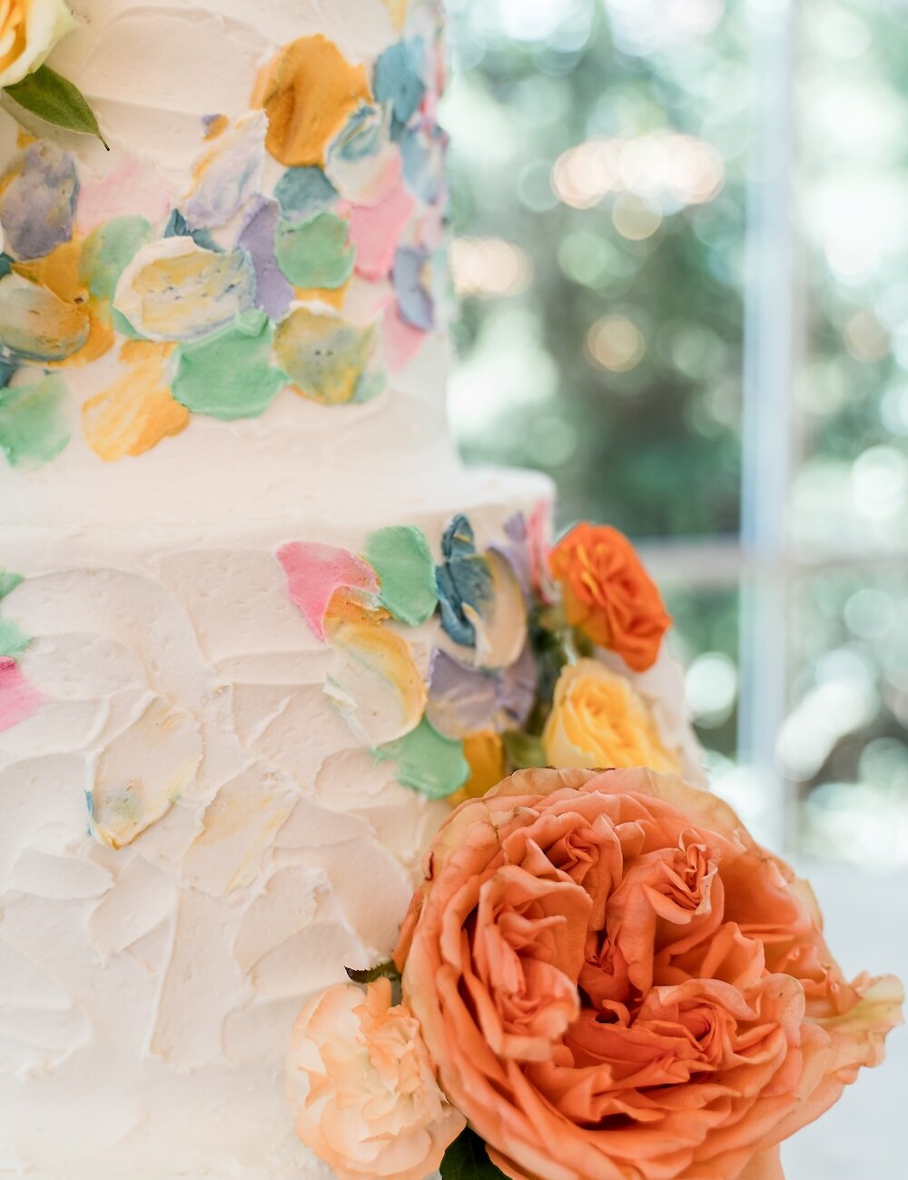 Close-up of a wedding cake featuring white textured buttercream, vibrant multi-colored buttercream swipes, and a large orange garden rose decoration.