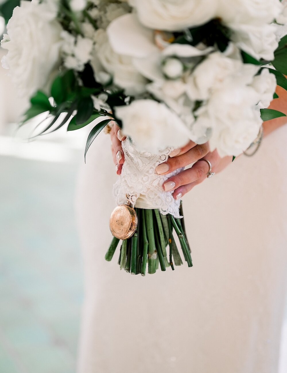 A close-up of a bridal bouquet of white roses and ranunculus, wrapped in lace and ribbon, with a vintage gold locket charm hanging from the stems.
