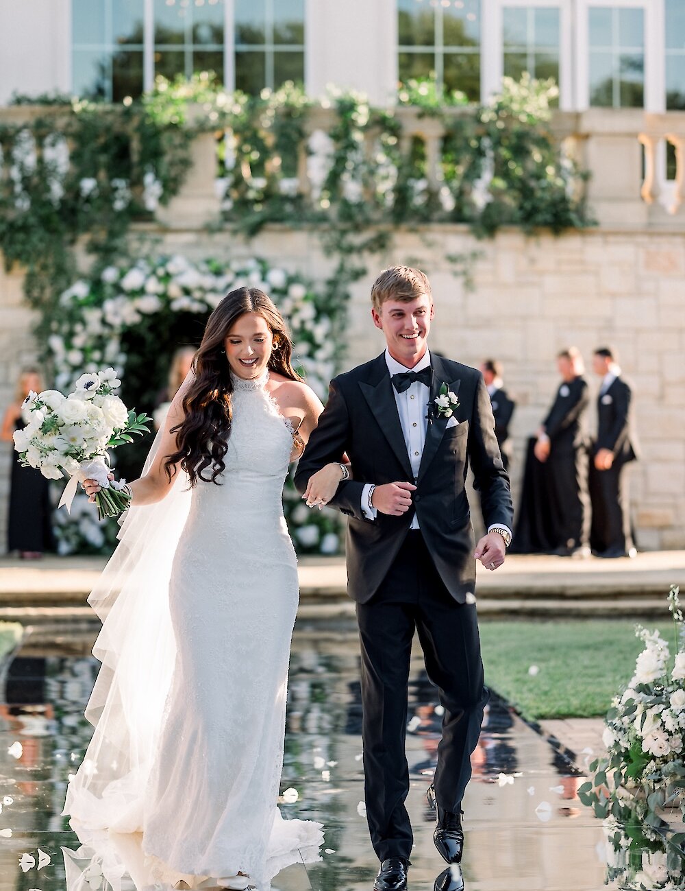 The smiling bride and groom walk hand-in-hand down a highly reflective surface, surrounded by flowers, with a stone building and archway visible in the background.