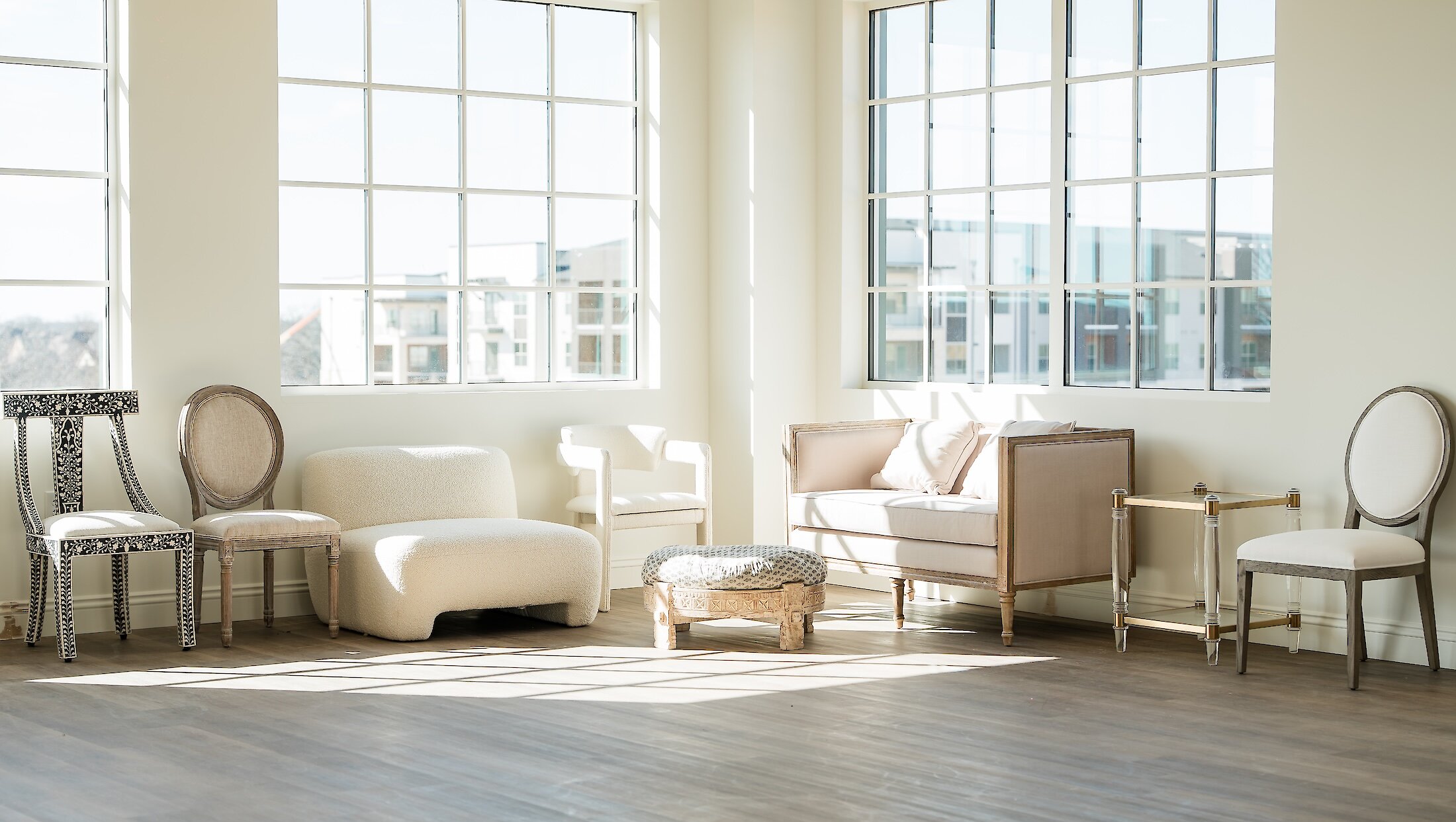 A sunlit, empty bridal suite or lounge area featuring a collection of eclectic white and beige furniture pieces set against large paneled windows.