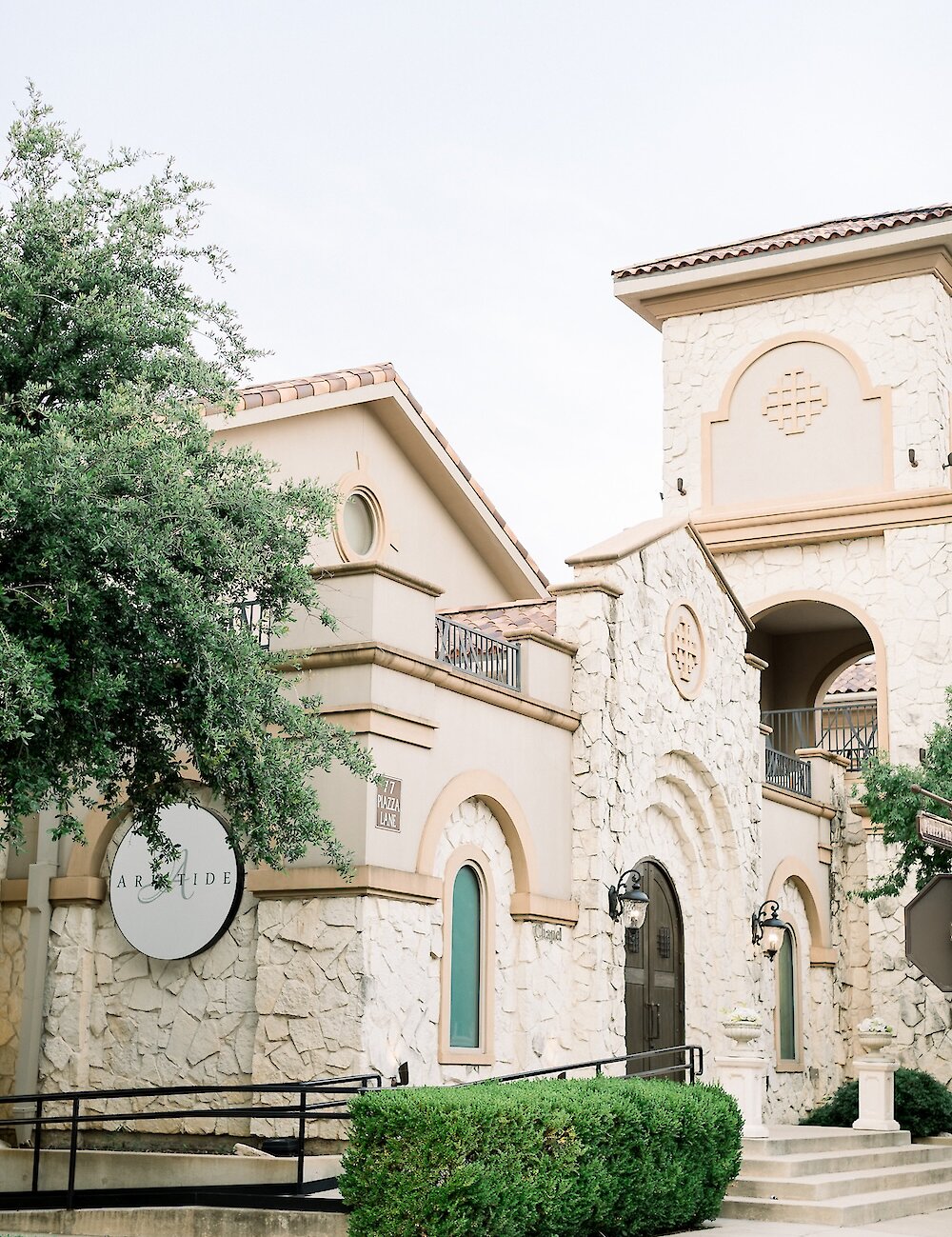 The exterior of a Mediterranean-style wedding venue featuring tan stone, terracotta roof tiles, arched entryways, and a circular sign.