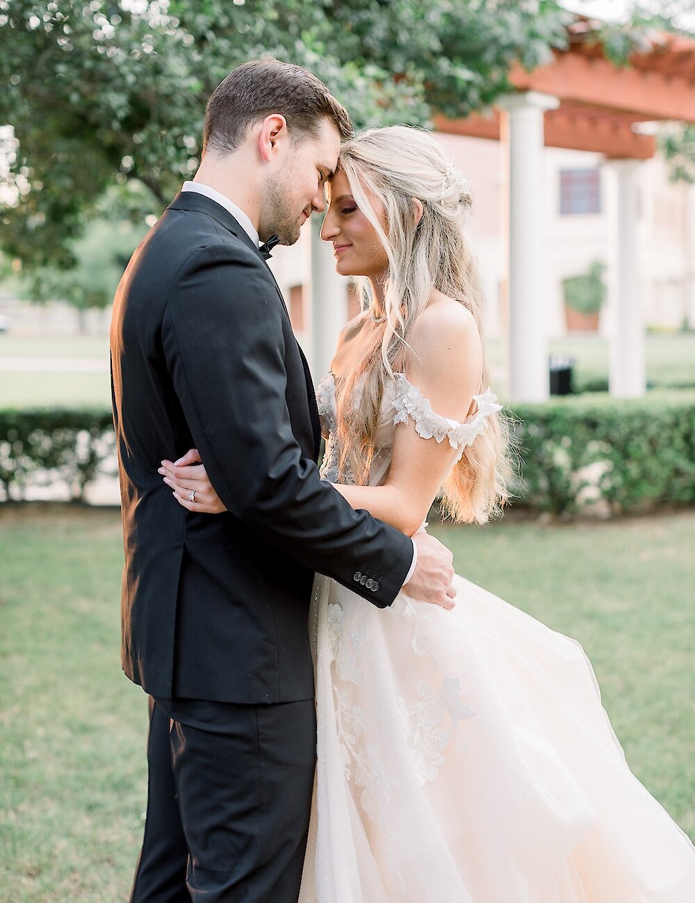 The bride and groom embrace and touch foreheads during a romantic outdoor portrait, with the groom wearing a black tuxedo and the bride in a ruffled off-the-shoulder gown.