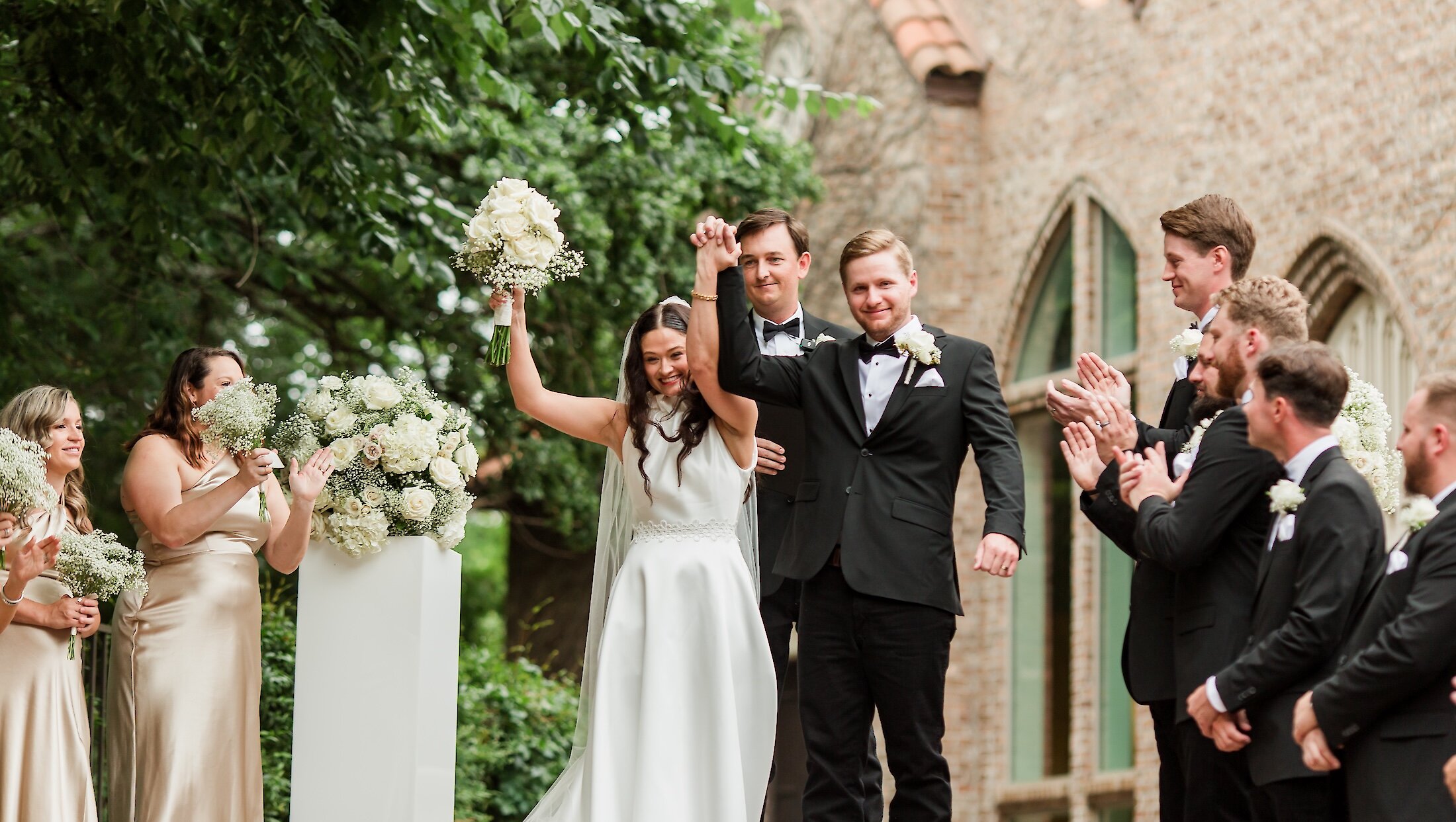 The elated bride and groom cheer and raise clasped hands after the ceremony, surrounded by their bridesmaids in champagne dresses and groomsmen in tuxedos.