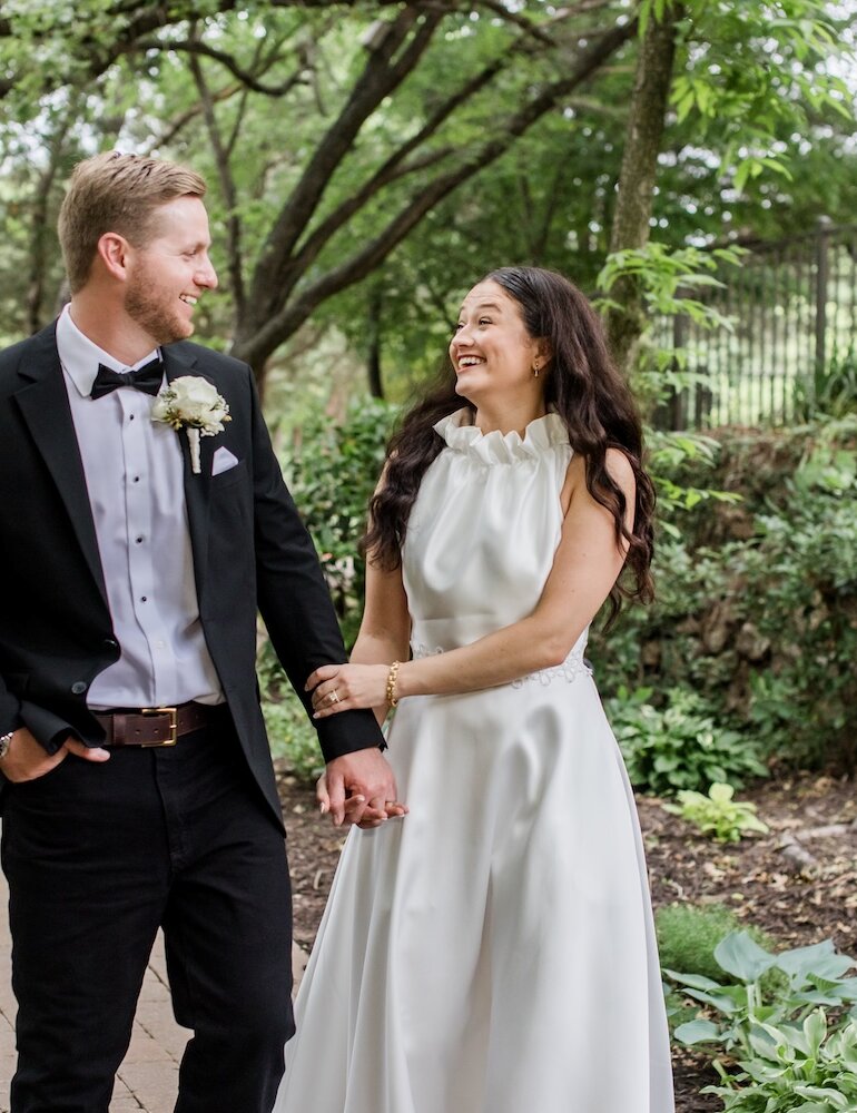 The smiling bride in a high-neck white gown and the groom in a tuxedo walk hand-in-hand down a brick pathway through a lush wooded garden.