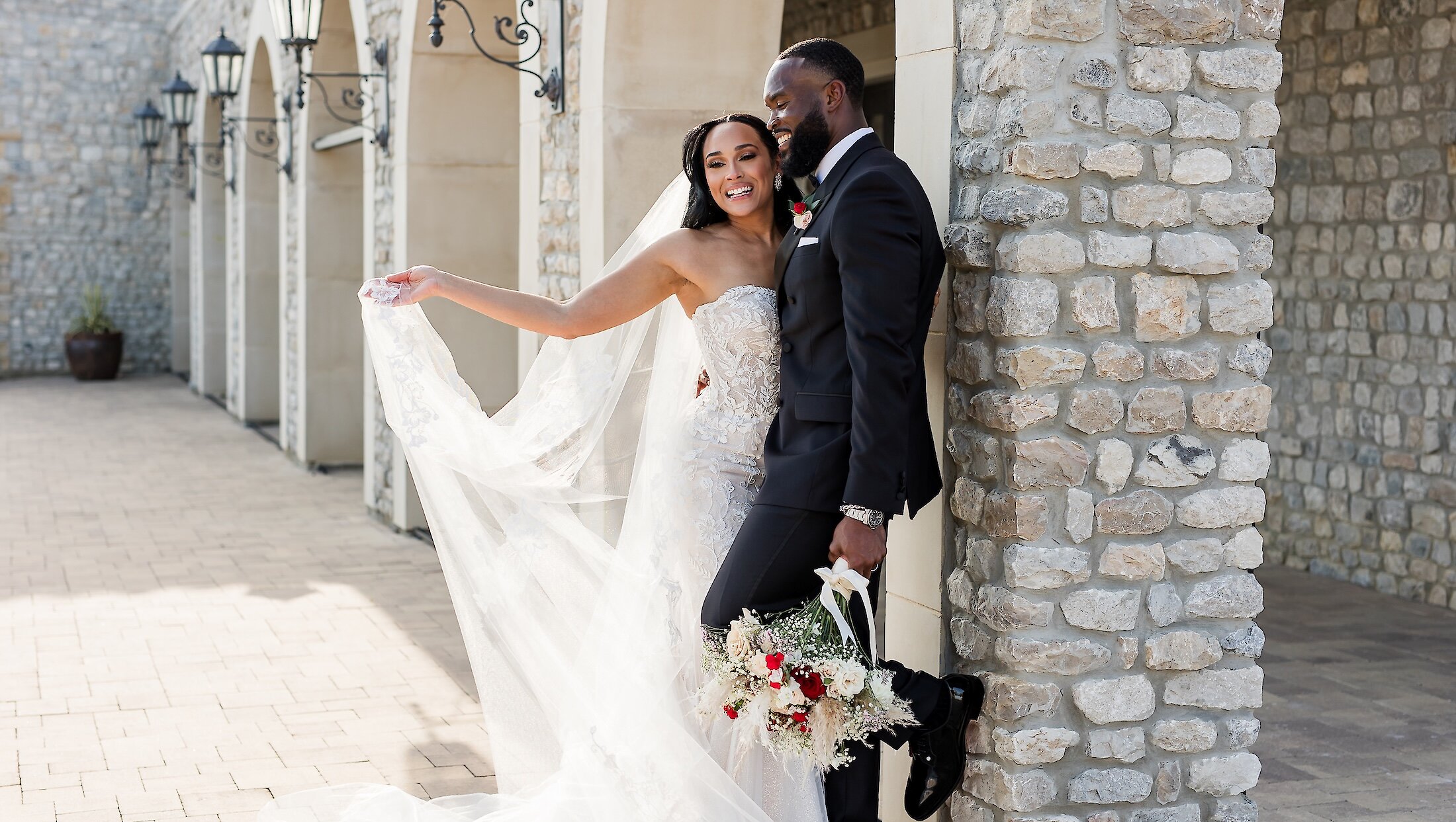 The bride holds her flowing veil while embracing the groom, who is leaning against a stone pillar with a bouquet, under a series of outdoor stone archways.