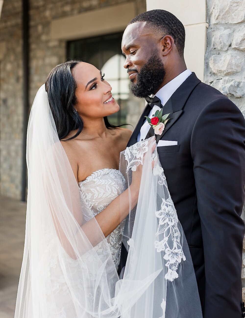 Close-up portrait of the bride and groom looking lovingly at each other, with the bride holding her lace-edged veil and the groom wearing a classic black tuxedo.