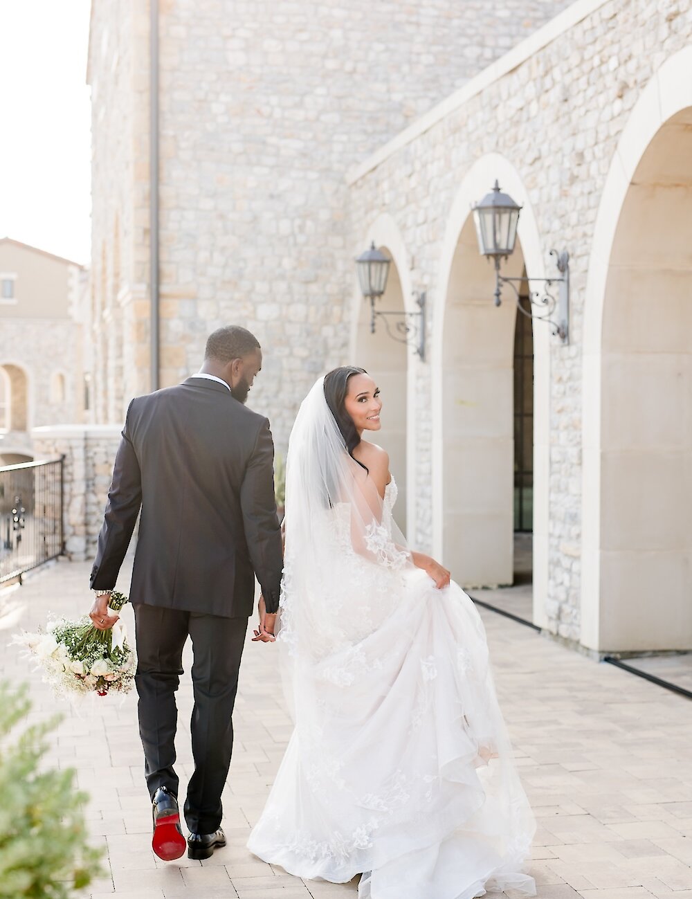 The bride smiles and looks back while walking away, holding hands with the groom, on a paved path outside a stone building with arched windows.