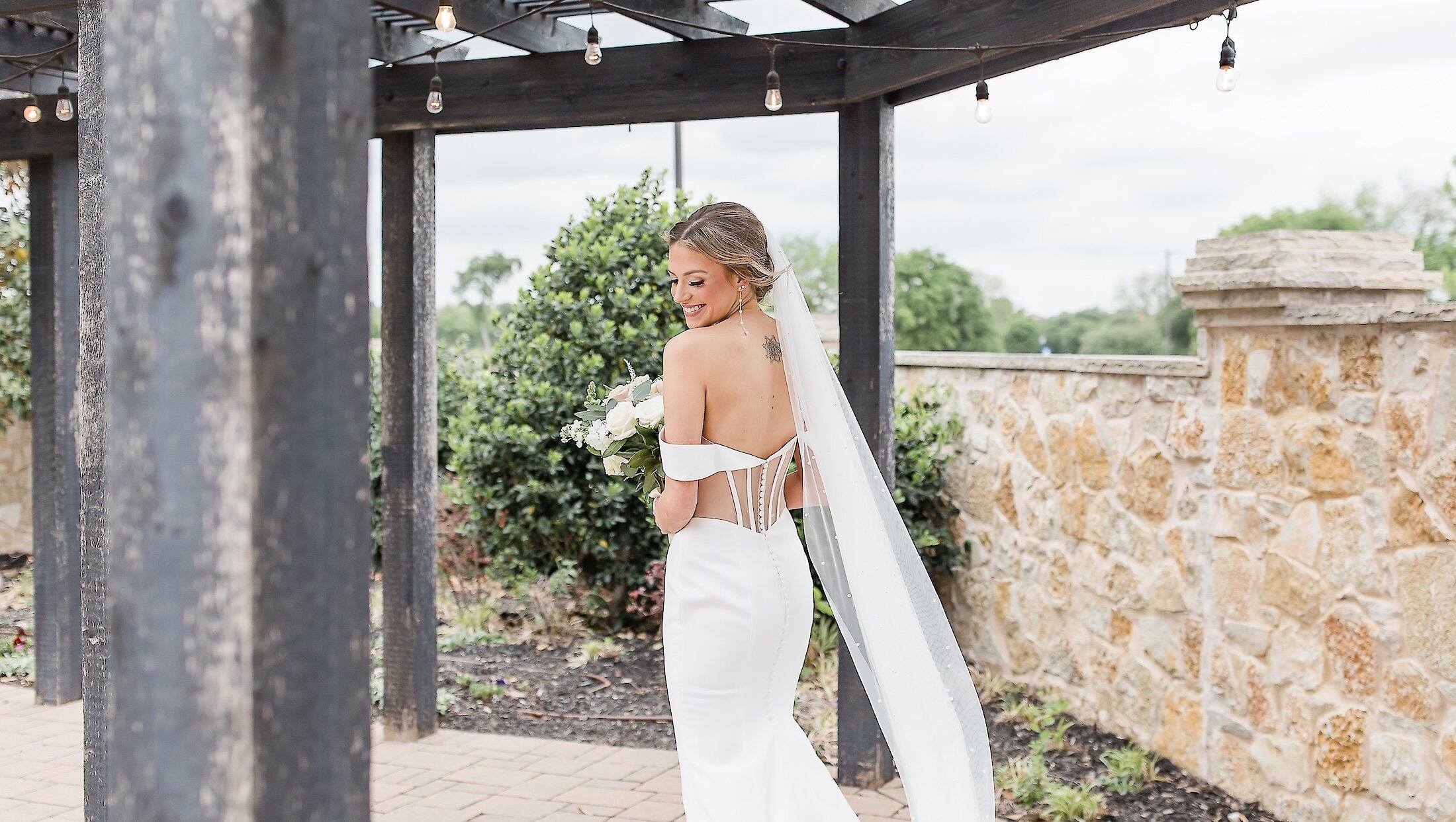 The bride smiles over her shoulder, showcasing her off-the-shoulder gown with visible boning, long veil, and white bouquet under a dark wood pergola.