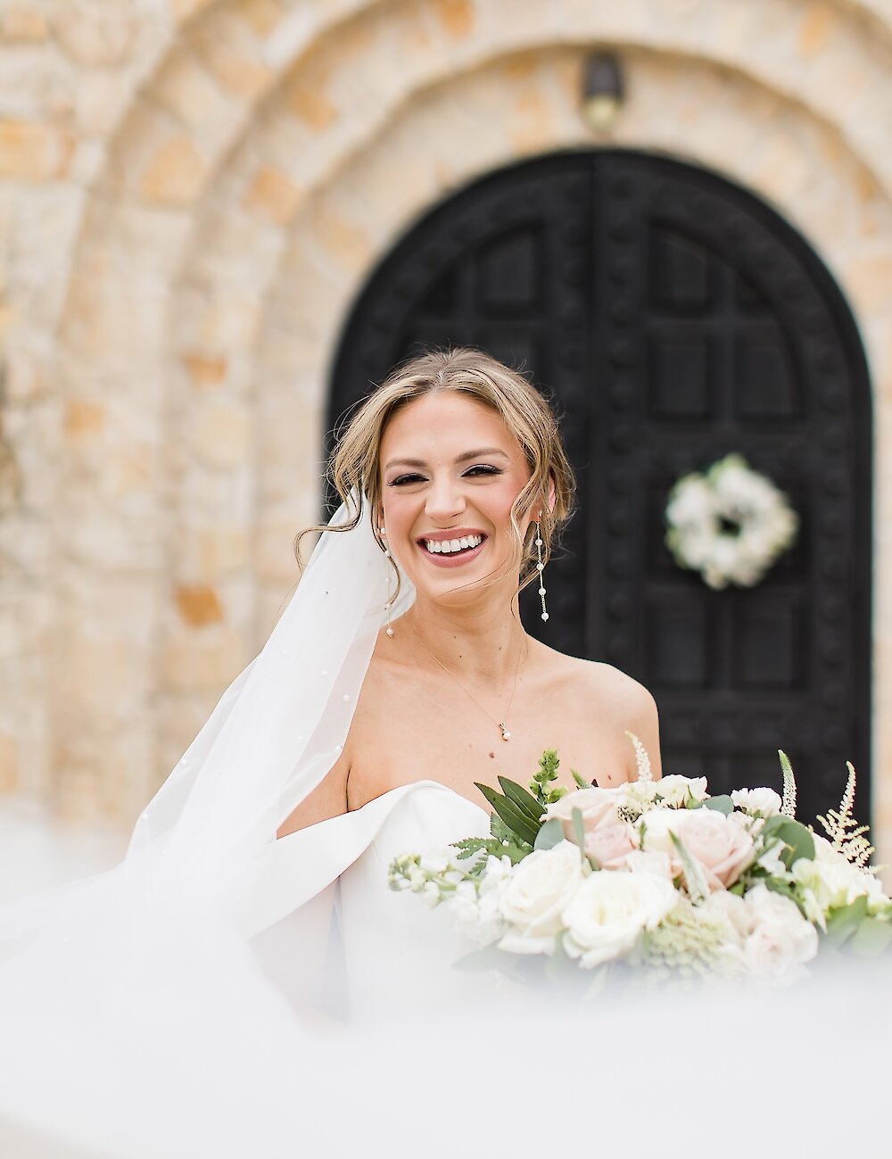 A close-up of the laughing bride holding her blush and white bouquet, with her veil flowing, posing in front of a dark arched chapel entrance.