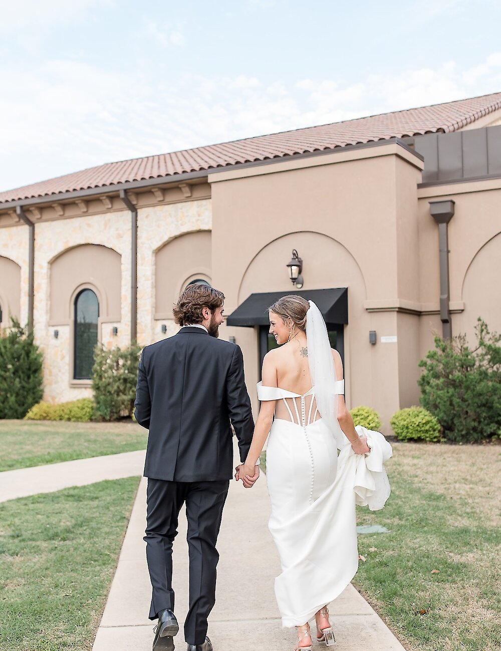 The bride looks back and smiles while walking away, holding hands with the groom, on a concrete path outside a tan Mediterranean-style wedding venue.
