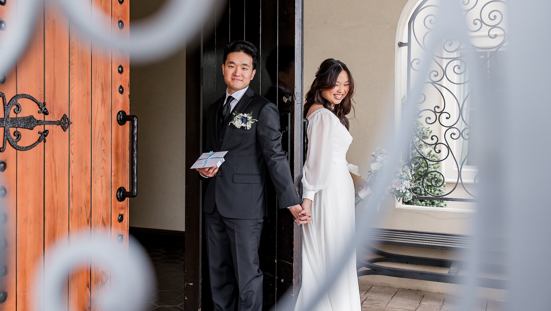 The smiling bride and groom hold hands while standing on opposite sides of a doorway for a first touch, with the groom holding a small card.
