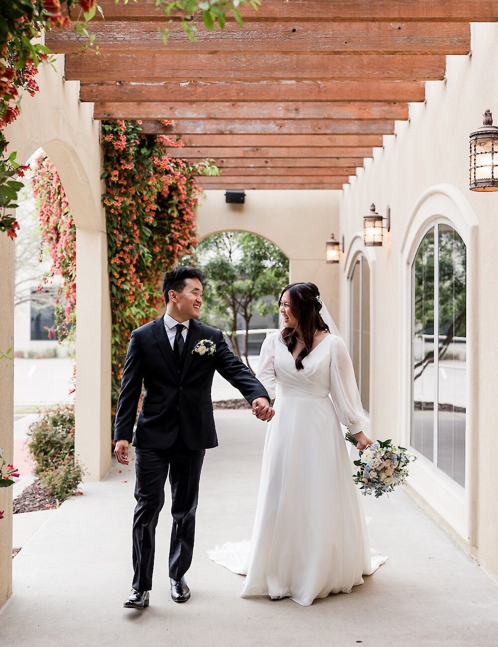 The bride and groom smile and hold hands while walking down a covered outdoor patio with wooden beams, arched openings, and red flowering vines.