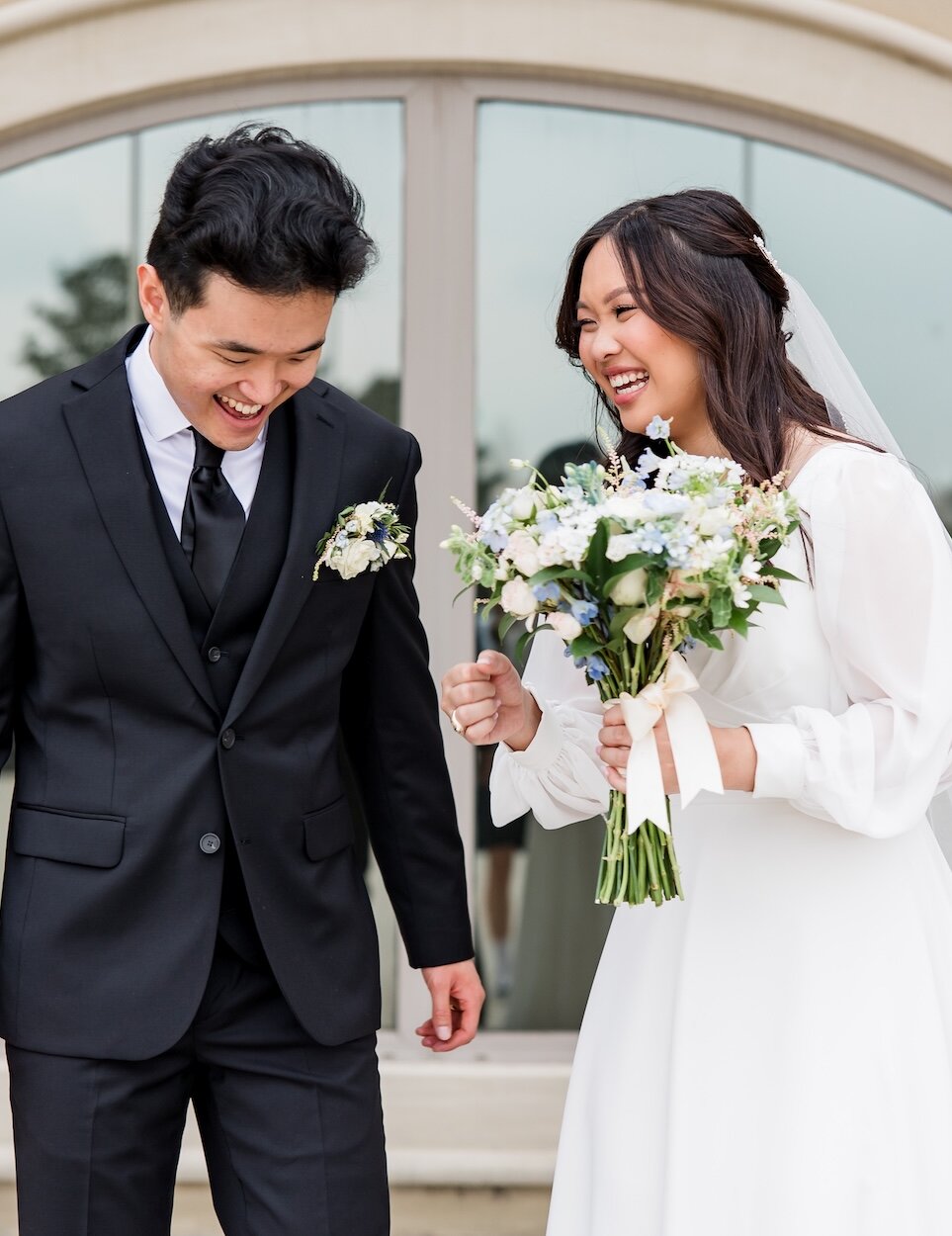 The bride and groom laugh and look down, with the bride holding a white and blue bouquet, posing outdoors in front of a tan arch window.