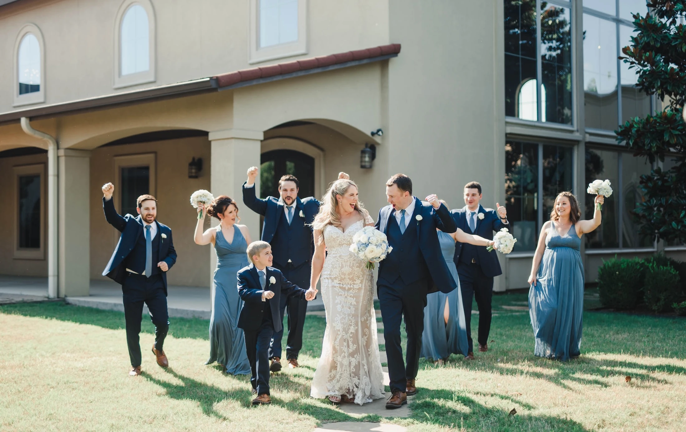The ecstatic bride and groom walk hand-in-hand while the wedding party cheers and raises their fists outside a venue on a sunny lawn.