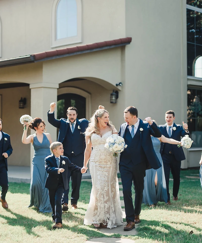 The ecstatic bride and groom walk hand-in-hand while the wedding party cheers and raises their fists outside a venue on a sunny lawn.