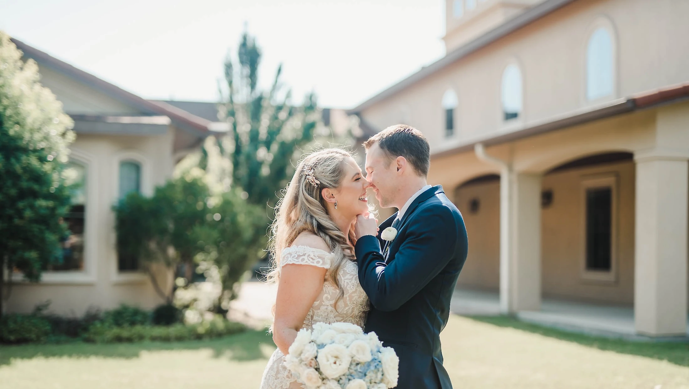 The bride and groom lean in to kiss while holding a white and blue bouquet, standing outdoors on a green lawn with a wedding venue building behind them.