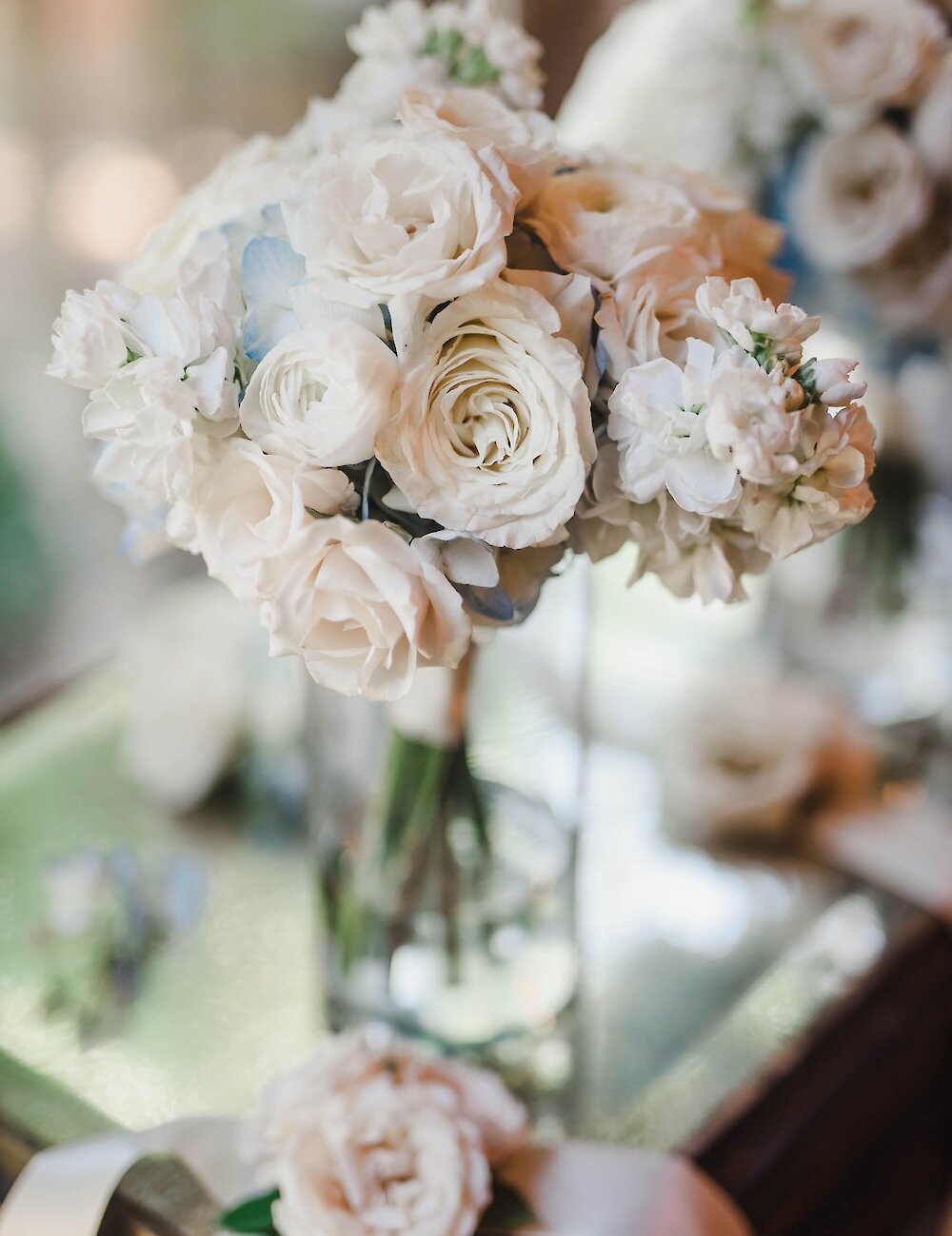 A beautiful arrangement of white and pale pink roses and hydrangeas stands in a glass vase on a mirrored surface, ready for the wedding ceremony.