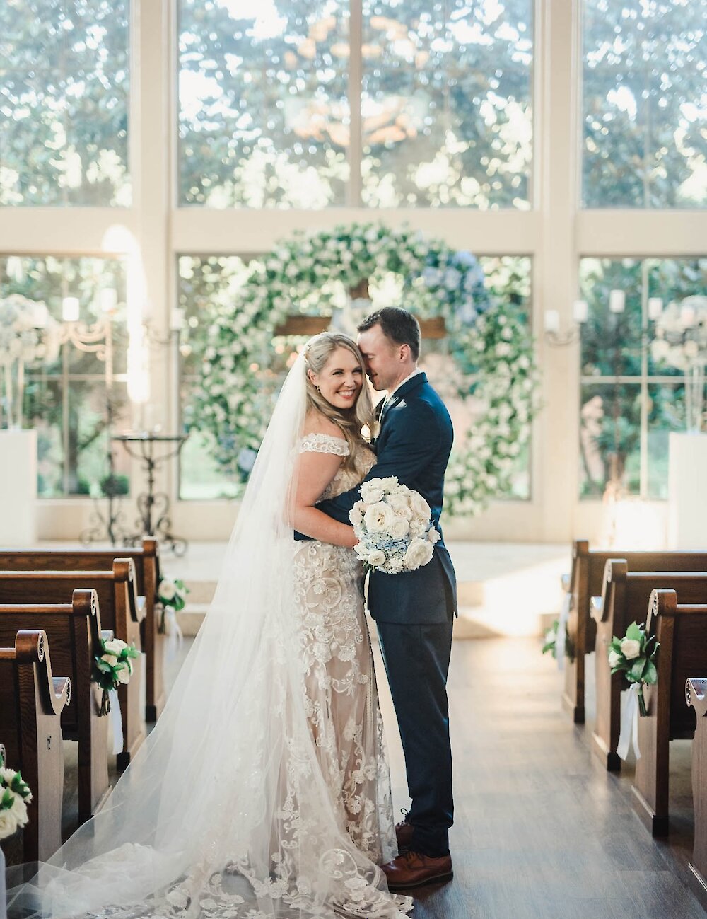 The bride and groom embrace and smile inside a chapel, standing in the aisle with pews on either side and a large floral wreath altar in the background.